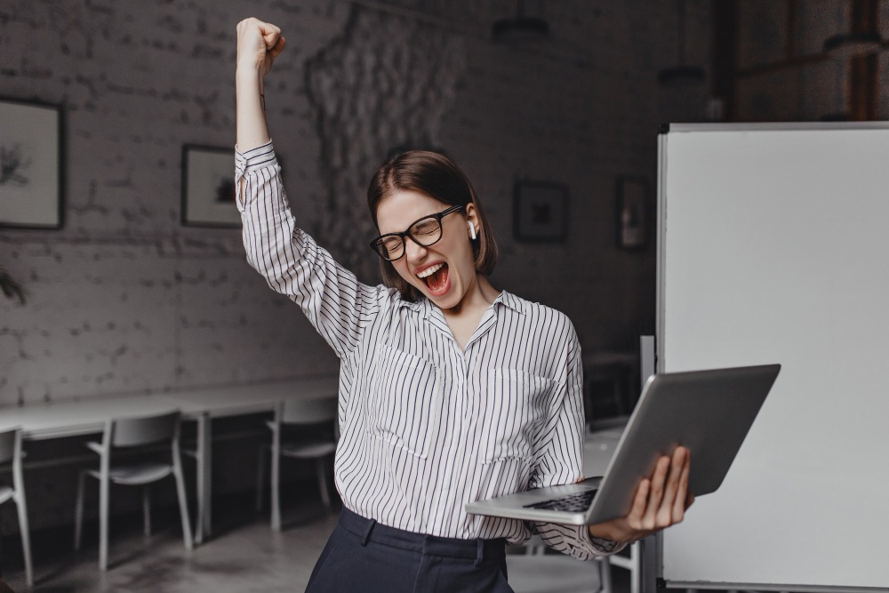 business-woman-with-laptop-in-hand-is-happy-with-success-portrait-of-woman-in-glasses-and-striped-blouse-enthusiastically-screaming-and-making-winning-gesture (1)
